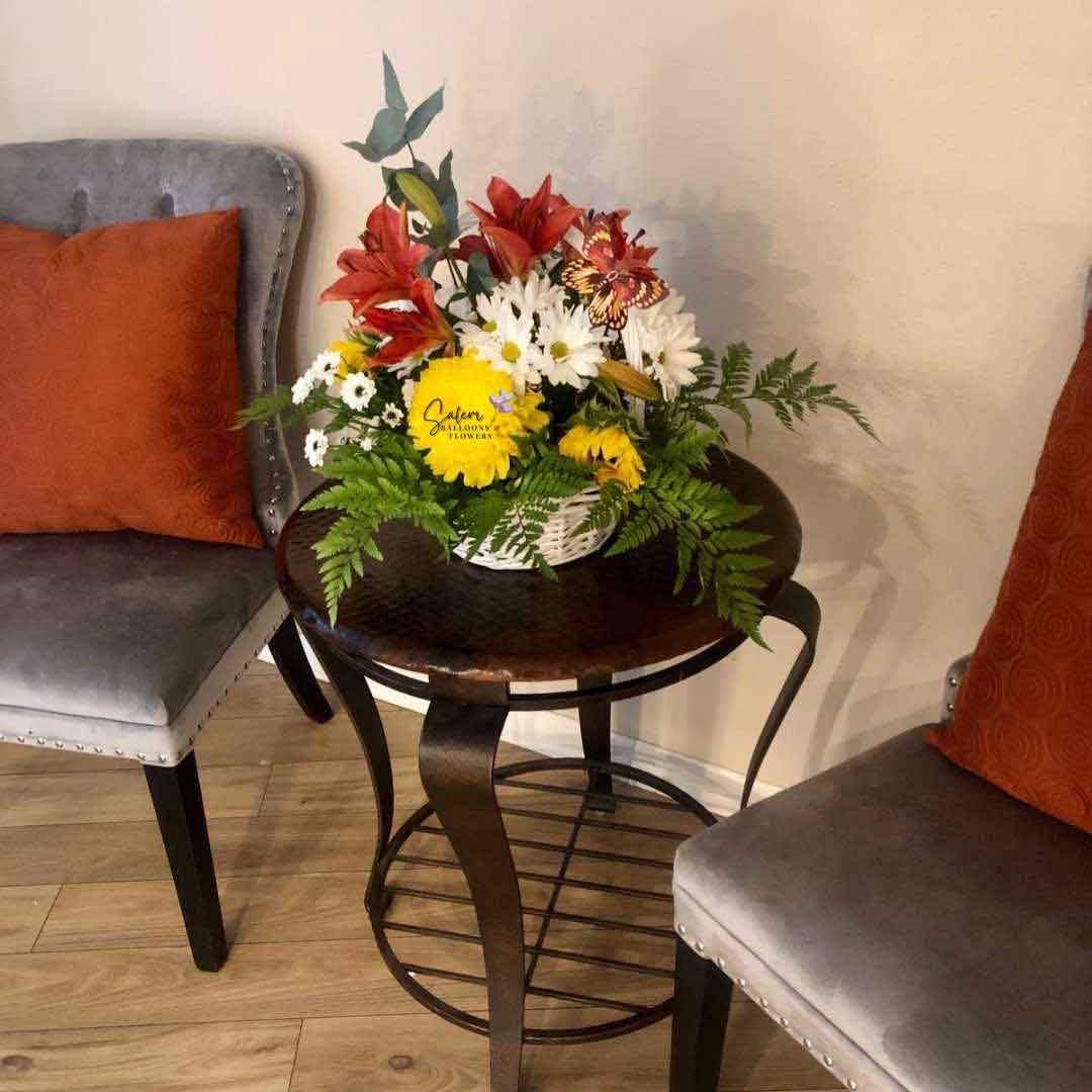 Seasonal flower basket with an array of beautiful flowers decorated with butterflies sitting on a center table between 2 chairs. Salem Flower delivery.