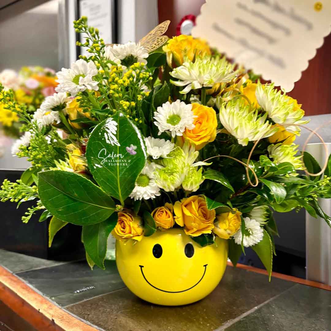 Smiley-face yellow vase with yellow spray roses and white flowers and greenery. Decorated with golden twicks and butterfly. Salem Oregon Flower Delivery.