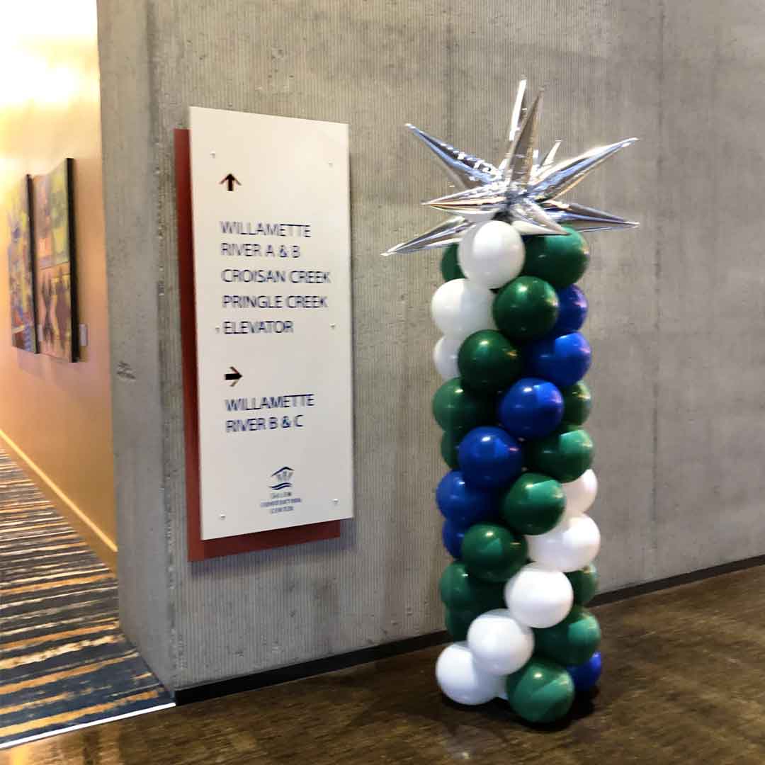 A Classic balloon column with a silver starburst balloon topper. The body of the balloon column comes in a swirl of green, blue, and white colors. Picture taken at the Willamette ballroom at The Salem Convention Center. Salem Oregon Balloon decorations.