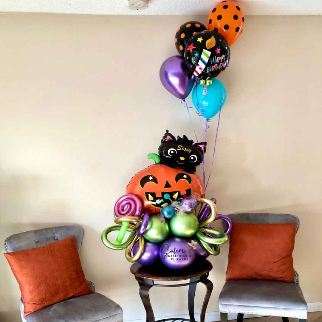 A Halloween themed balloon bouquet, featuring a pumpkin balloon with a cute black cat, decorated with helium balloons and curly balloons, customized with name standing on a table between 2 chairs. Oregon Balloon delivery