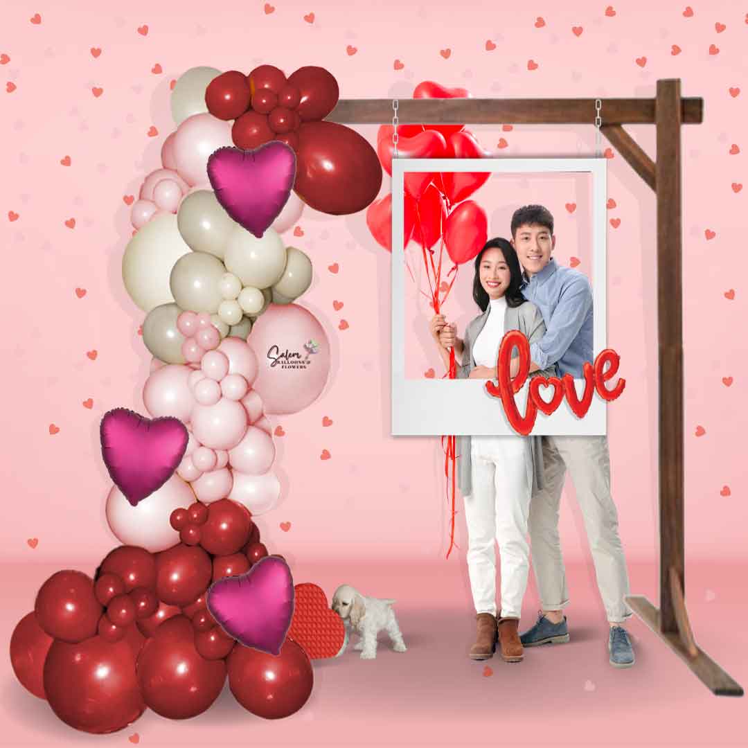 A Couple holding balloons and posing underneath a Valentine's Day Balloon Decoration. Decorated with a charming balloon garland on a rustic wood frame and complete with a cute polaroid frame prop, with a LOVE sign balloon. Salem Oregon. Balloon decorations.