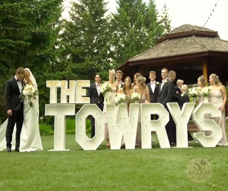Wedding setup with a large Marquee Letter display spelling "The Towrys" with the newly wedds standing at the left and their wedding company standing behind the marquee Letter display on an outdoors venue by Salem Balloons and Flowers.