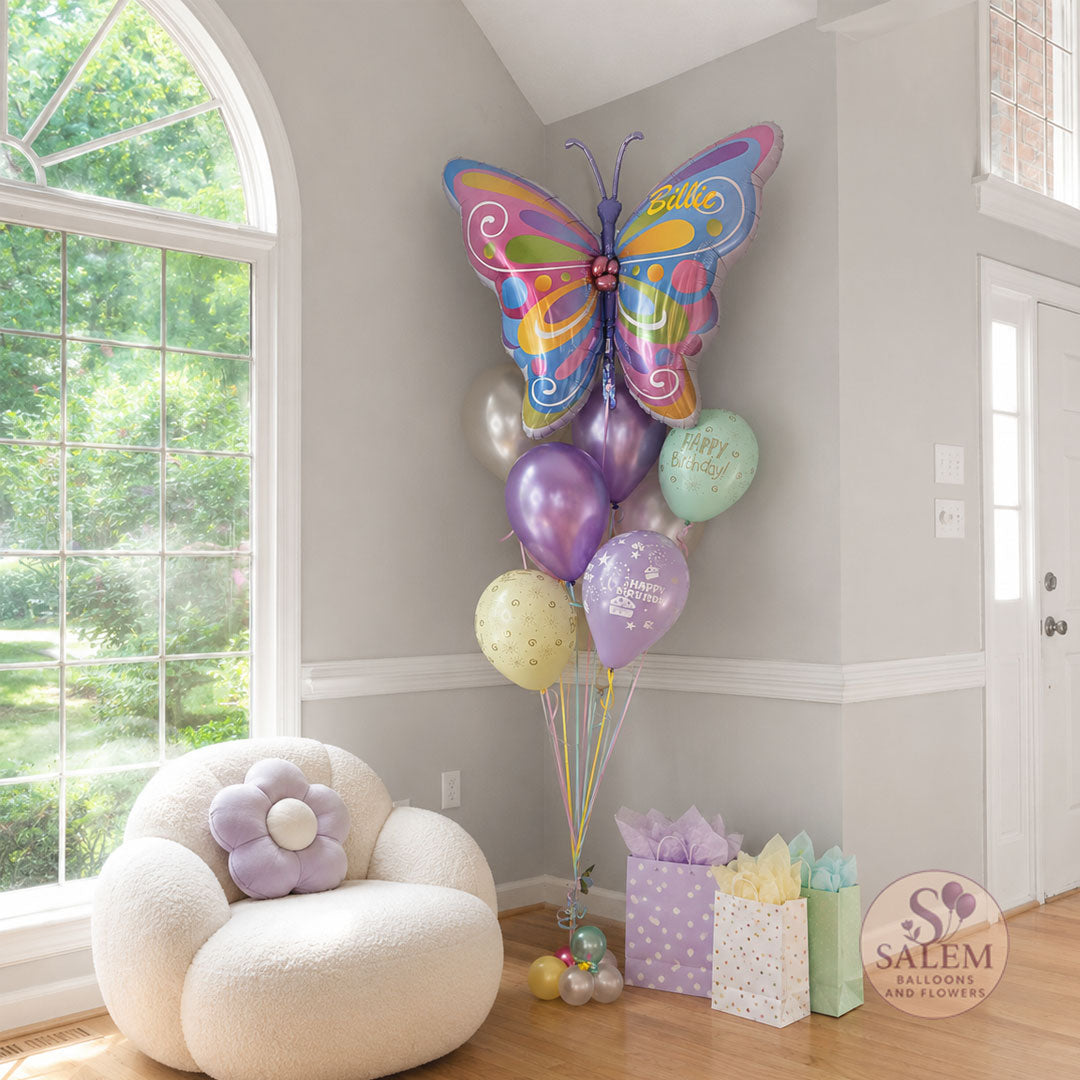 A whimsical pastel butterfly birthday balloon bouquet displayed in a bright luxury home entryway with tall arched windows, hardwood floors, and soft neutral walls. The colorful helium balloon arrangement is styled in the corner beside pastel gift bags and a cream boucle cloud chair. Creating an elegant and joyful birthday celebration scene with crisp natural lighting. with a Salem Balloons and Flowers logo. Balloon delivery Willamette Valley.