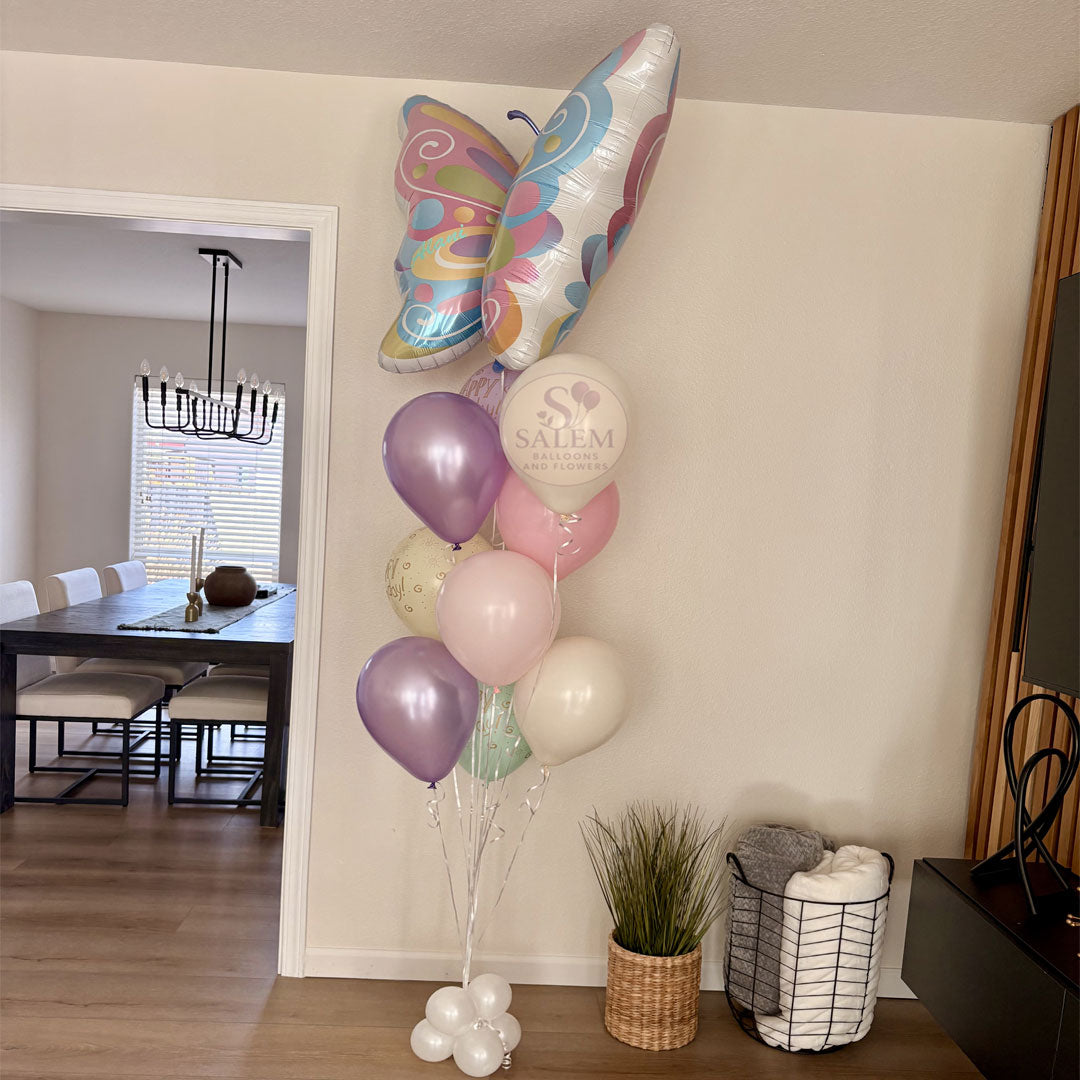 A whimsical pastel butterfly birthday balloon bouquet displayed in home living room. The colorful helium balloon arrangement is styled in the corner with a Salem Balloons and Flowers logo. Balloon delivery Willamette Valley.