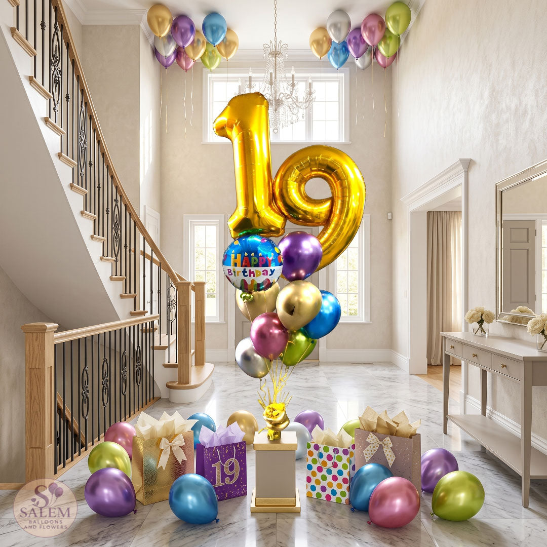 A colorful 19th birthday balloon bouquet featuring metallic gold number balloons, a "Happy Birthday" foil balloon, and chrome-tone latex balloons centered in a bright, modern foyer with marble flooring, a grand staircase, ceiling balloons, and gift bags arranged below. locally delivered by Salem Balloons and Flowers.