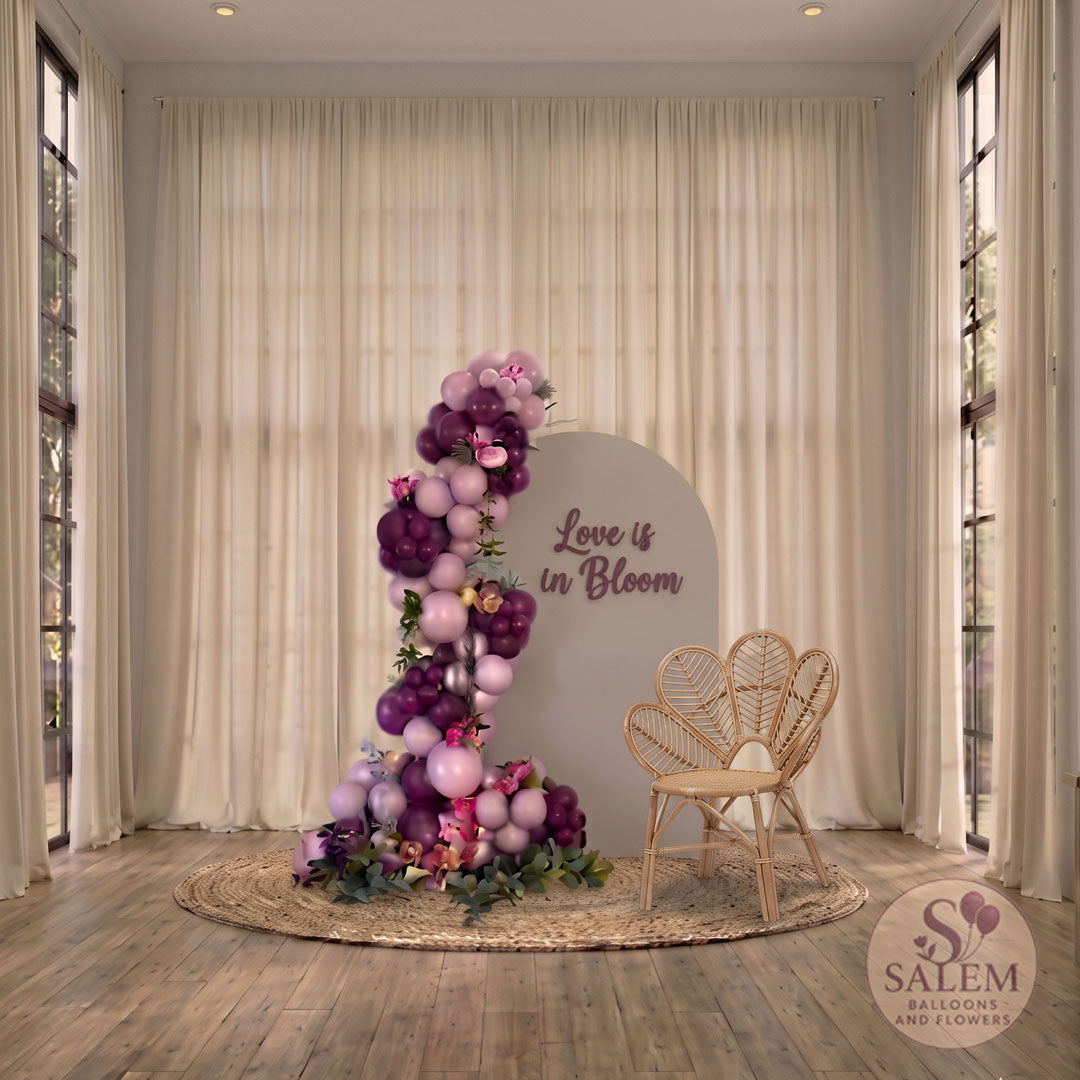 Plum and purple organic balloon garland with flowers next to a "Love is Bloom" backdrop and rattan chair, featuring luxury balloon decor by Salem Balloons and Flowers in Salem, Oregon and the Willamette Valley.