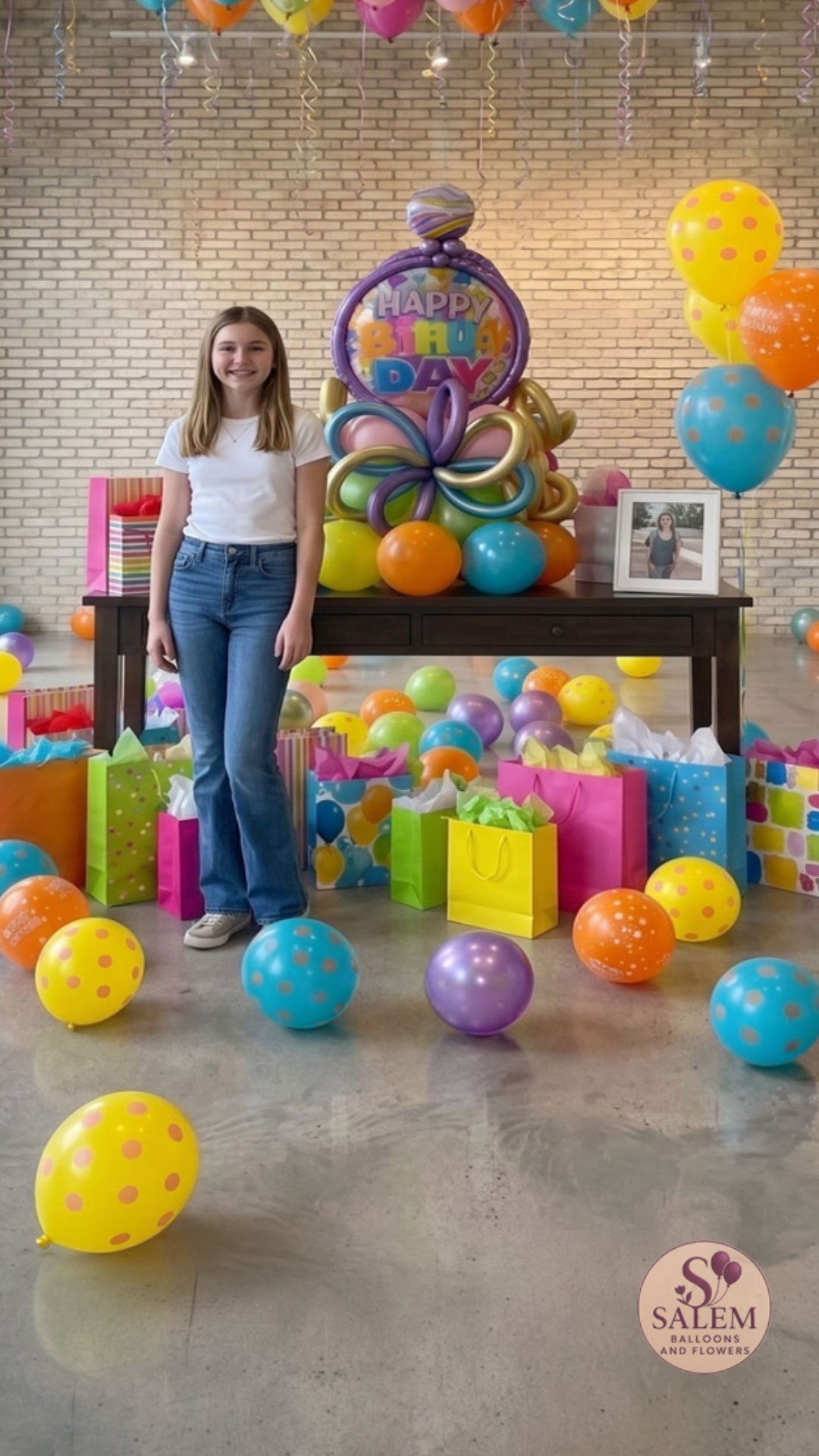 A young teenage girl in a white t-shirt and jeans stands next to a large, colorful 'Happy Birthday' balloon bouquet setup on a dark wood table in a bright room with scattered floor balloons and a full ceiling of helium balloons. Birthday balloon delivery in Salem Oregon by Salem Balloons and flowers.