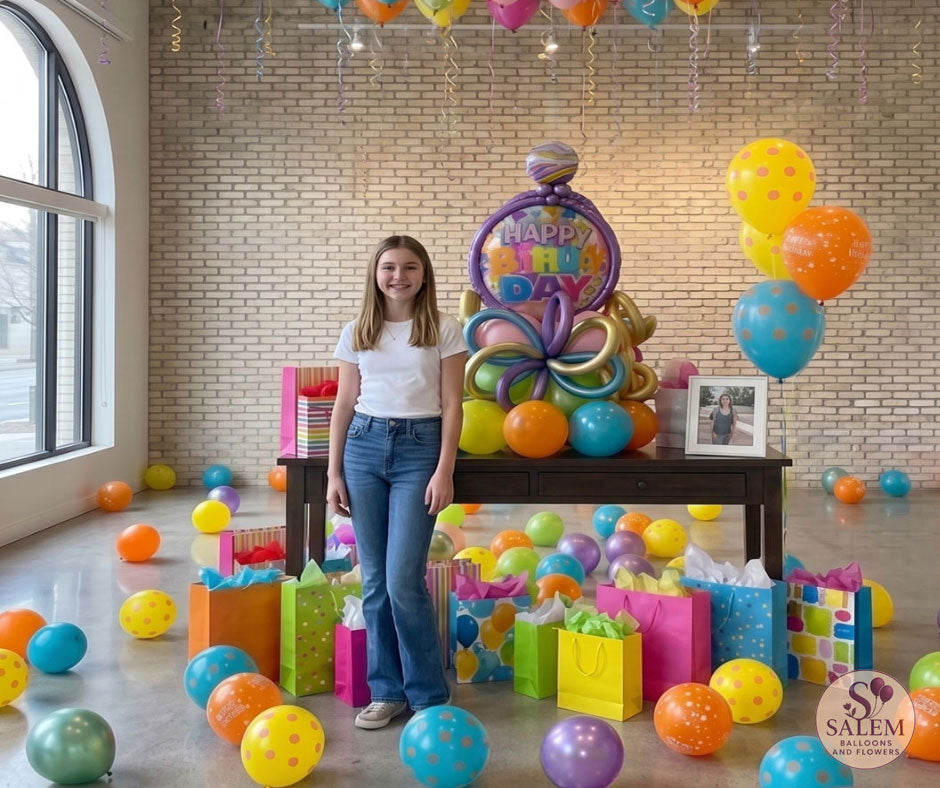 A young teenage girl in a white t-shirt and jeans stands next to a large, colorful 'Happy Birthday' balloon bouquet setup on a dark wood table in a bright room with scattered floor balloons and a full ceiling of helium balloons. Birthday balloon delivery in Salem Oregon by Salem Balloons and flowers.