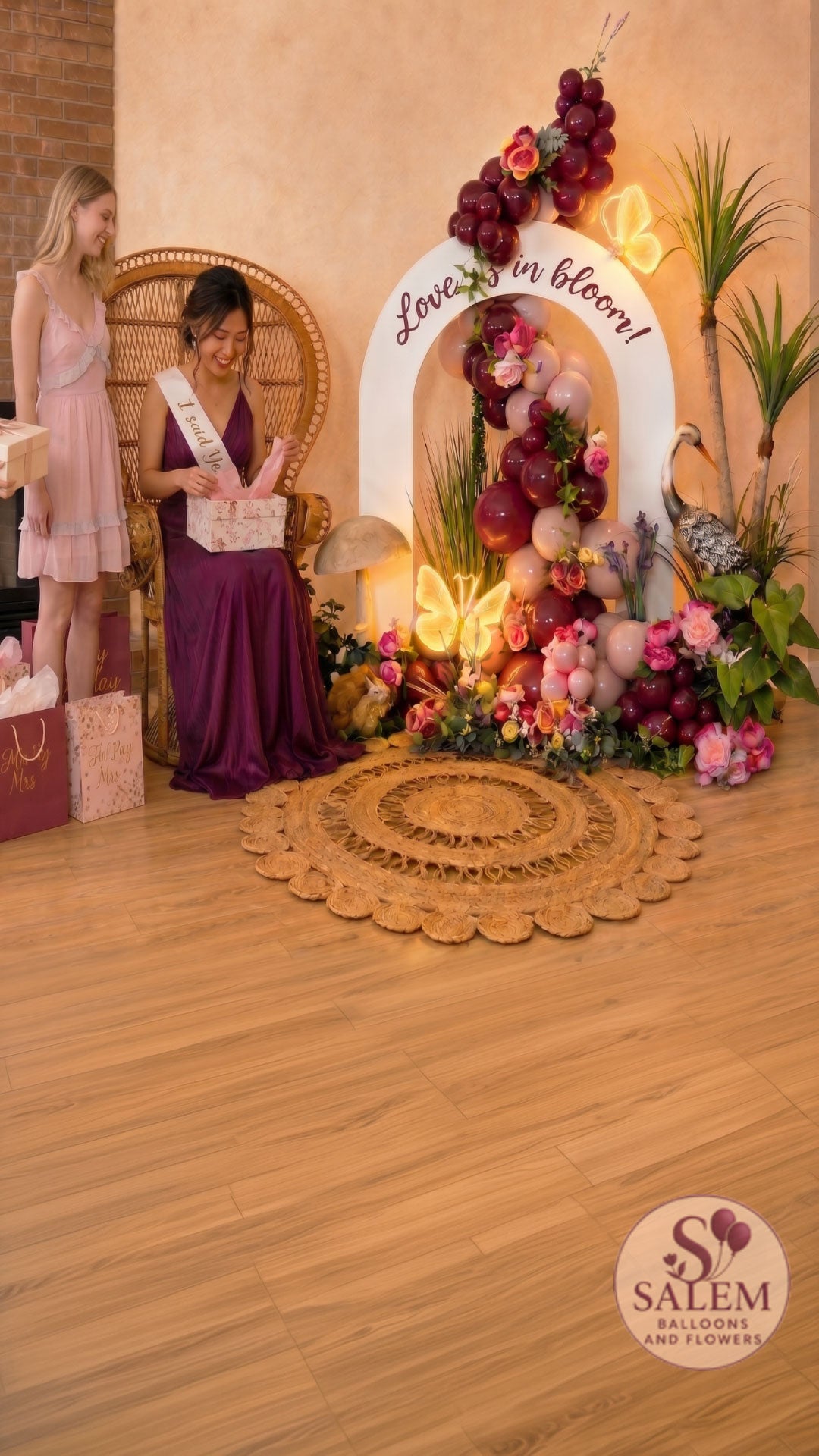 Luxury bridal shower balloon décor featuring a bride-to-be opening gifts beside the Blooming Signature Moment floral balloon installation with peacock chair, illuminated butterfly lights, luxury balloon garland, and premium event styling in burgundy and pink by Salem Balloons & Flowers in Salem, Oregon.