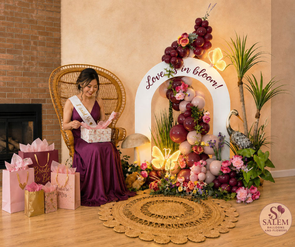 Luxury bridal shower balloon décor featuring a bride-to-be opening gifts beside the Blooming Signature Moment floral balloon installation with peacock chair, illuminated butterfly lights, luxury balloon garland, and premium event styling in burgundy and pink by Salem Balloons & Flowers in Salem, Oregon.