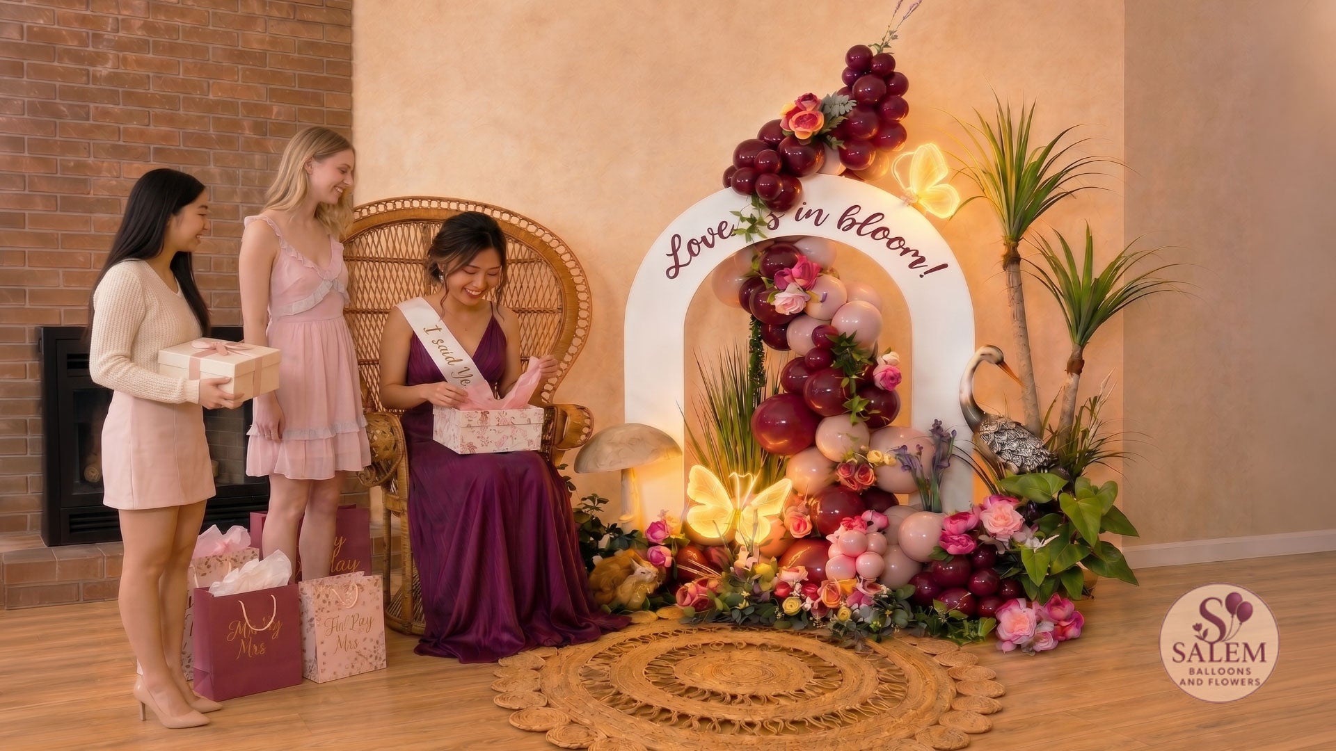 Luxury bridal shower balloon décor featuring a bride-to-be opening gifts beside the Blooming Signature Moment floral balloon installation with peacock chair, illuminated butterfly lights, luxury balloon garland, and premium event styling in burgundy and pink by Salem Balloons & Flowers in Salem, Oregon.