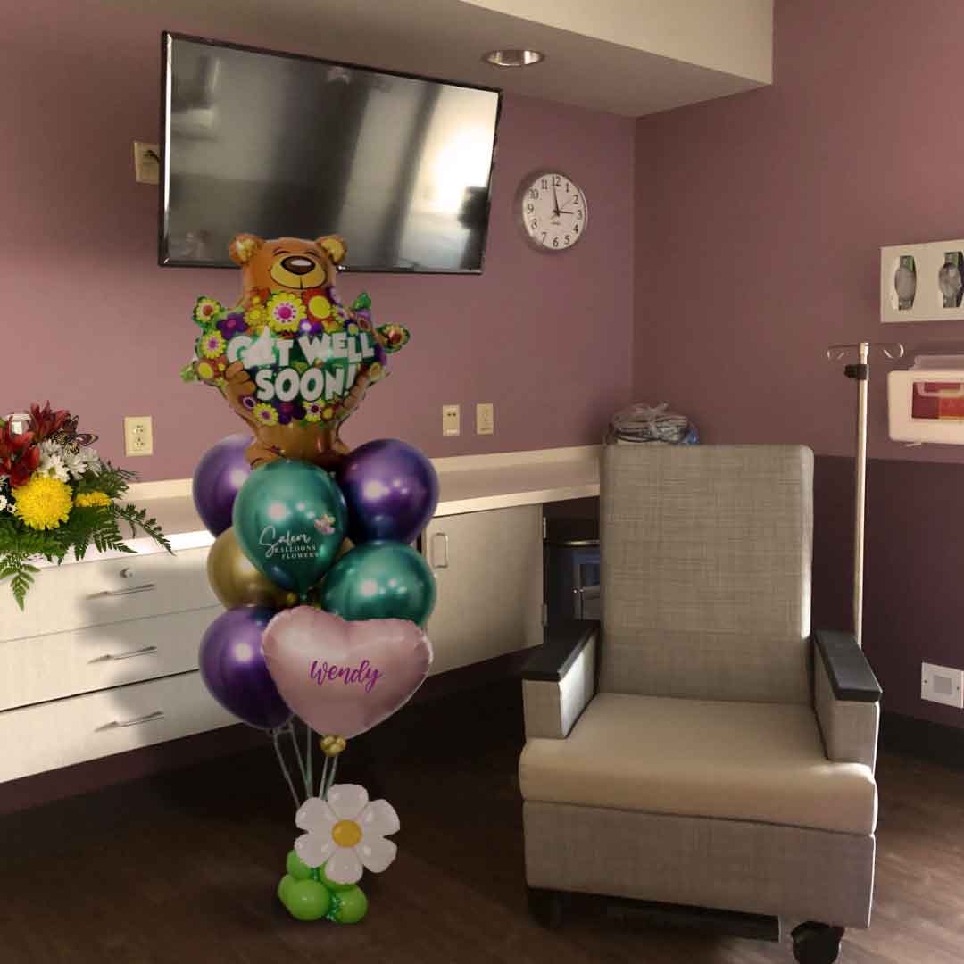 A hospital room with flowers and a Get Well helium balloon bouquet. Featuring a large teddy bear balloon holding flowers with a "Get Well" message in vibrant colors. Oregon Balloon delivery.