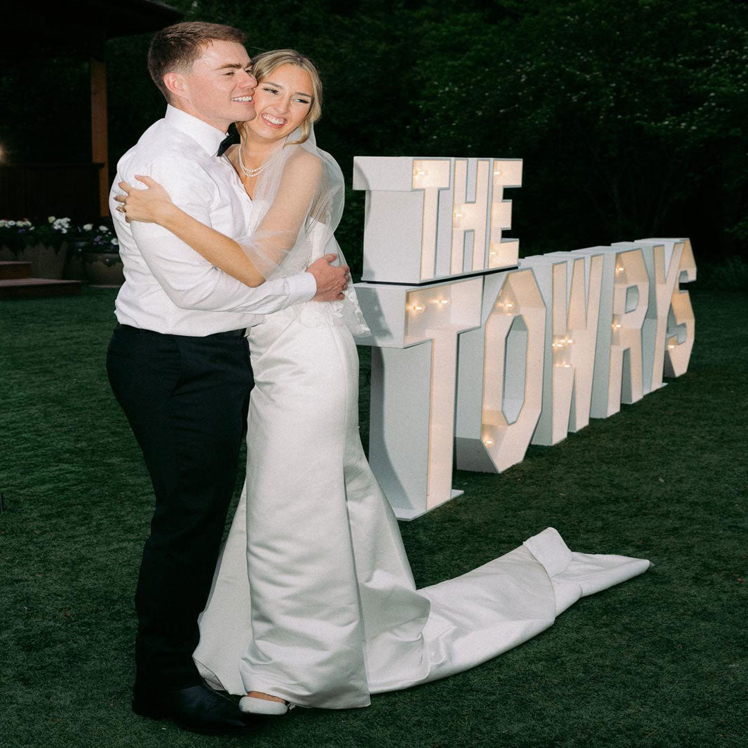 Bride and groom embracing beside glowing “THE TOWRYS” marquee letter display during an outdoor Oregon wedding reception. Elegant wedding focal installation by Salem Balloons and Flowers.