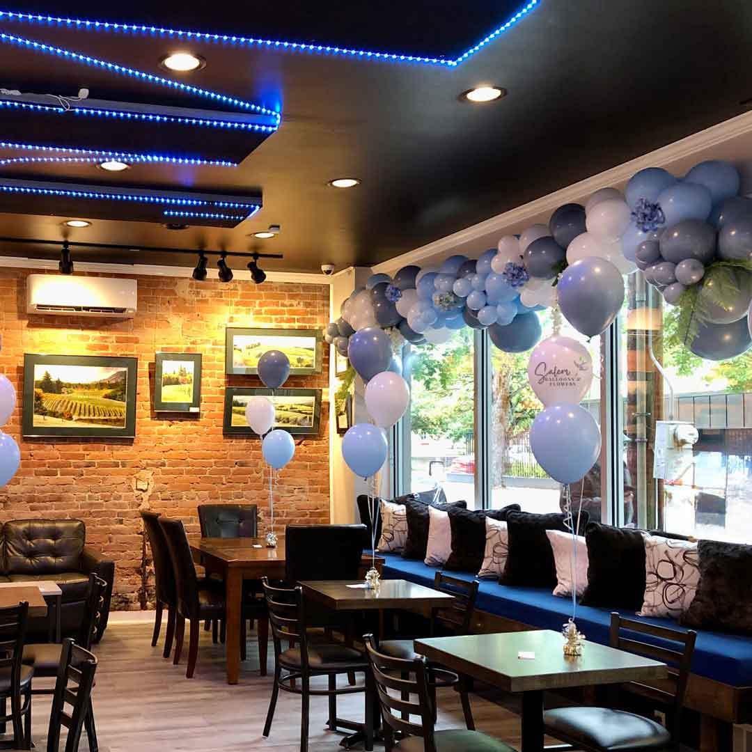 An organic balloon garland in shades of slate blue, steel blue, and white, embellished with floral accents and installed along the window seating at NW Cru. Designed by Salem Balloons and Flowers for a luxury wine tasting birthday celebration in McMinnville, Oregon.