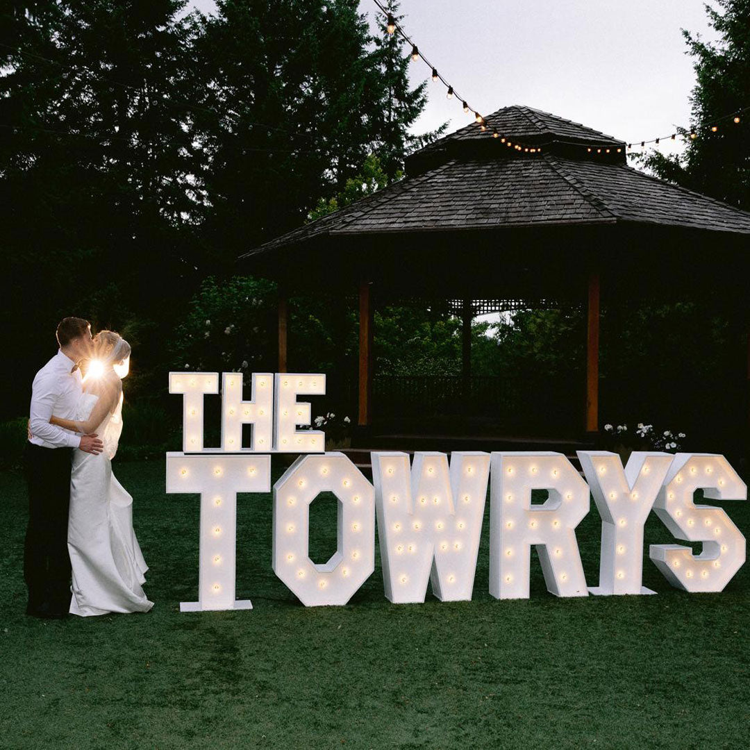 Bride and groom kissing beside glowing “THE TOWRYS” marquee letters under string lights at Paradise Springs wedding venue in Monroe, Oregon. Elegant outdoor wedding marquee display by Salem Balloons and Flowers.