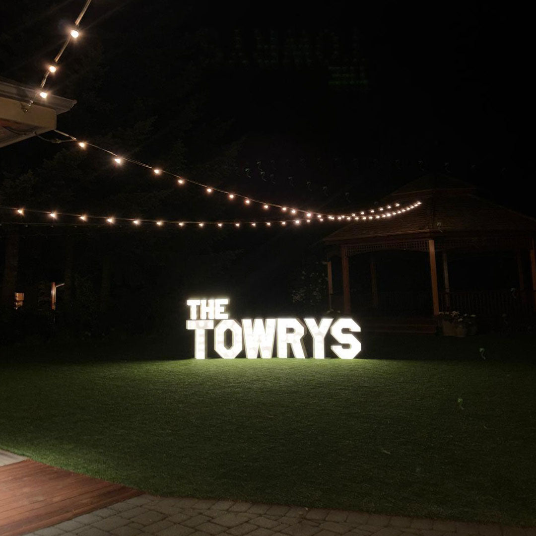 Illuminated “THE TOWRYS” wedding marquee letters glowing beneath string lights during a nighttime outdoor wedding reception in Monroe, Oregon. Luxury marquee installation by Salem Balloons and Flowers.