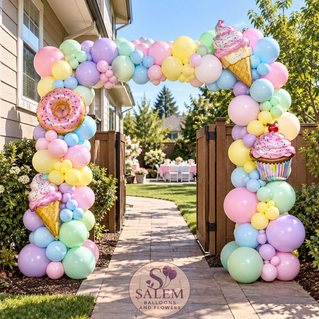 A pastel square balloon arch decoration set up outdoors on a backyard patio walkway, featuring large balloons in pink, yellow, blue, and green, with balloon donuts, ice cream cones, and a cupcake with a Salem Balloons and Flowers logo.